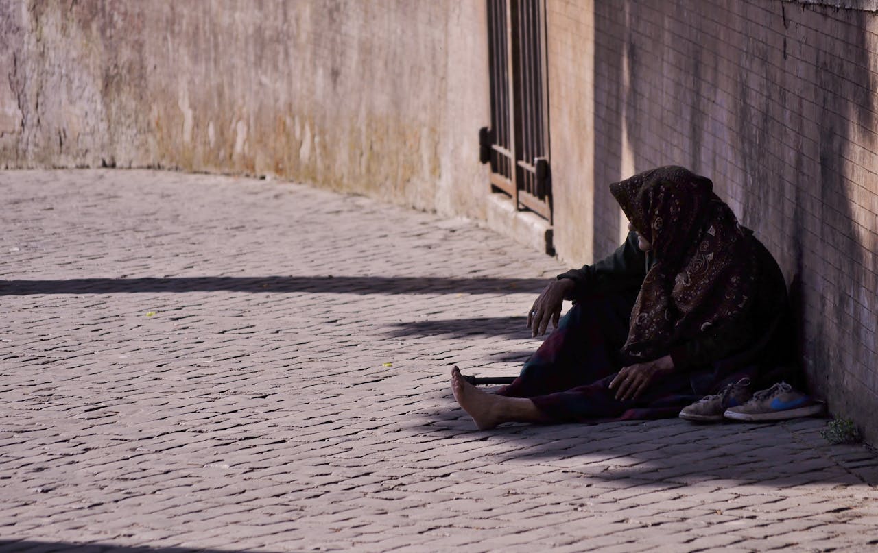 A woman sitting alone on a cobblestone street, covered with a headscarf.