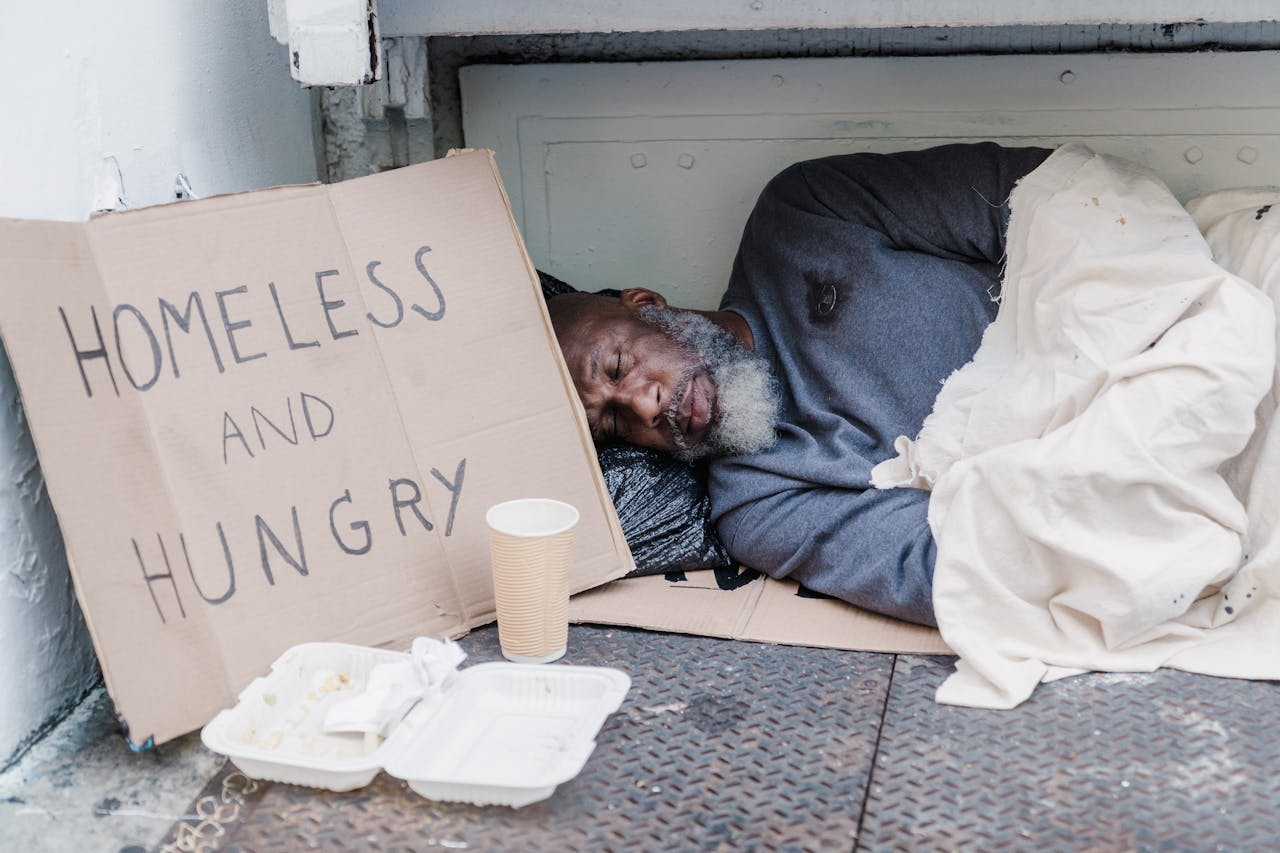 Elderly homeless man sleeping on the street with a sign reading 'Homeless and Hungry'.